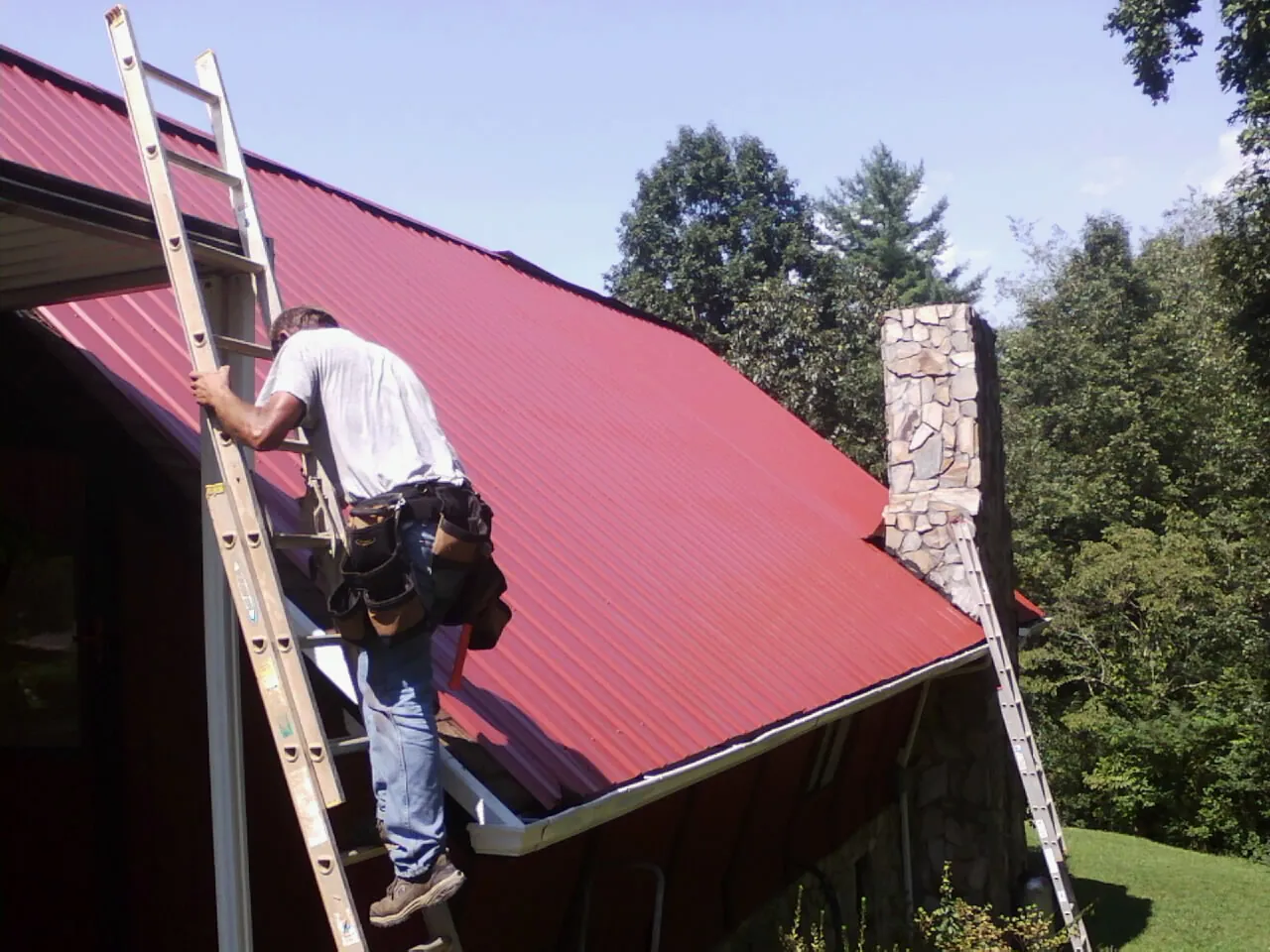 Roofer on a ladder beside a red metal roof and stone chimney — Soffit & Fascia Repair by Roofing Brothers in Woodruff